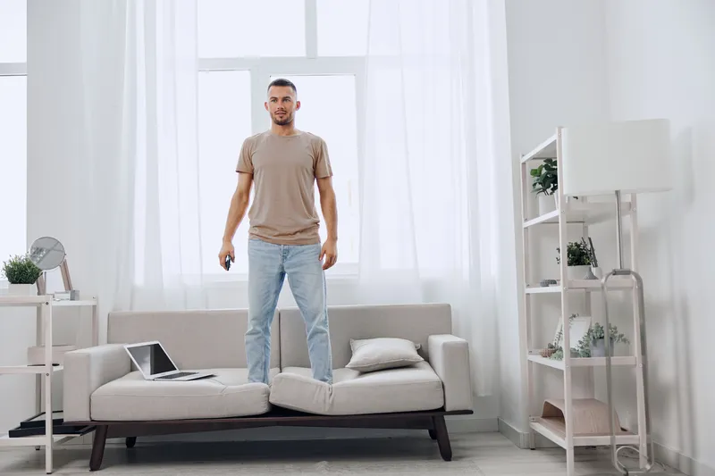 Happy man standing on couch, confidently using smartphone in modern, bright living room with minimalistic decor and soft colors, embracing the comfort of home.