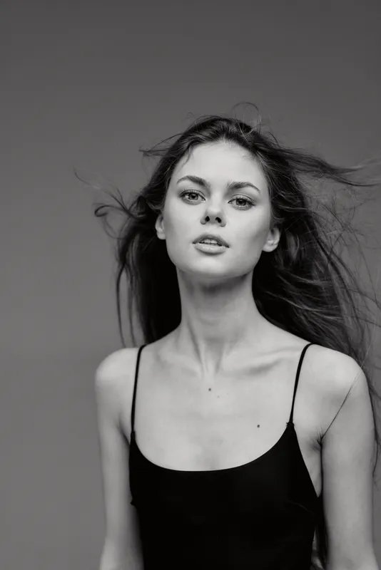 Black and white portrait of a young woman with loose hair and a minimalist black top, expressing calm and strength against a neutral background.