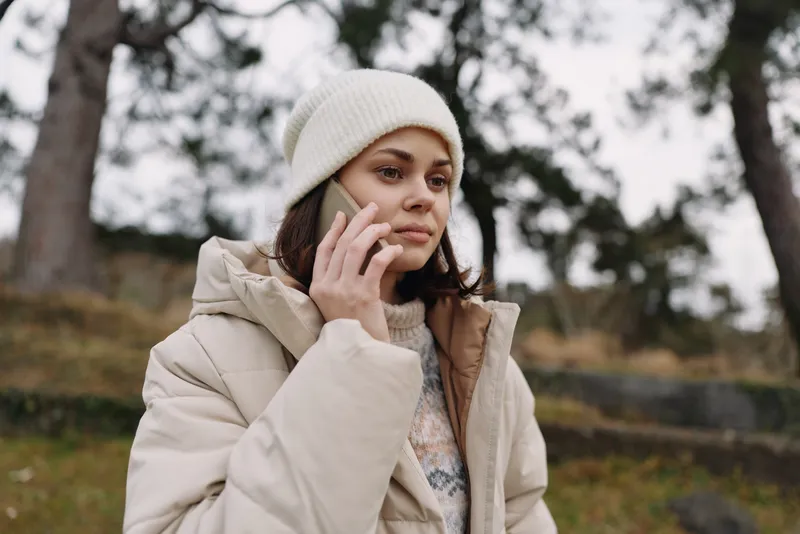 Woman in a light beige winter coat and knitted beanie speaks on a smartphone outdoors, standing in a park with trees and a cloudy sky, calm expression during a chilly day.