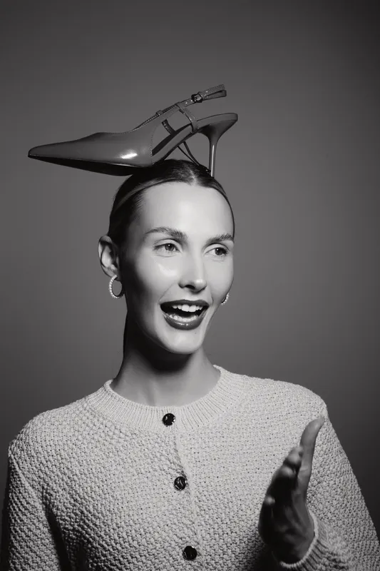 Smiling caucasian woman with light skin wears knitted sweater and high heel shoe balanced on head in studio. Concept of fashion, humor, and creative portrait with playful mood.