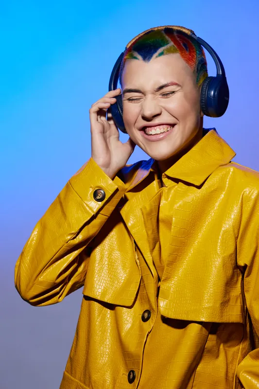 Young woman with colorful short hair wearing headphones and a vibrant yellow jacket enjoys music with a joyful expression against a bright blue background.