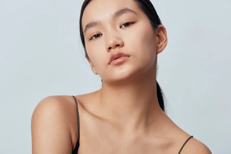 Portrait of young woman with natural skin and dark hair wearing black top against light background. Calm face and soft lighting for beauty concept and skincare model with minimal makeup.