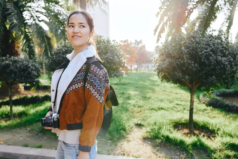 Photography of a woman with camera outdoors in a sunlit park, wearing a patterned jacket and scarf, capturing travel moments and natural light with calm confidence