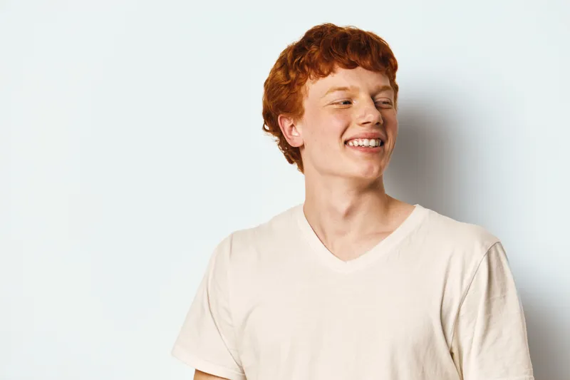 Young man with red hair and smile posing near white wall in casual light t shirt, looking sideways. Male portrait, youth happiness and casual fashion style in studio.