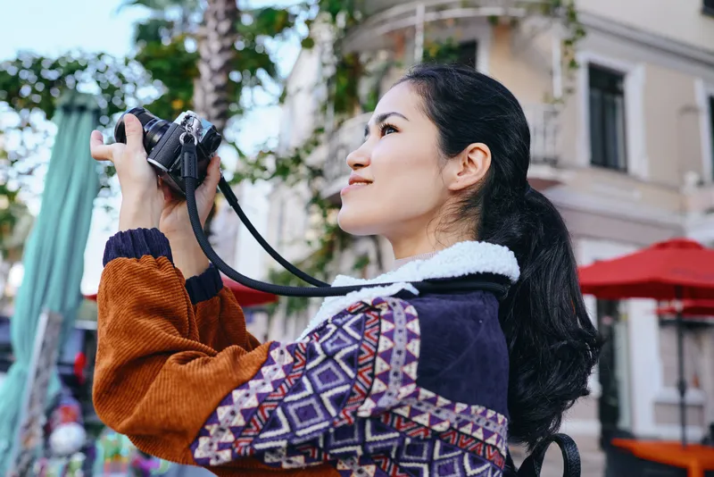 Photography of a woman holding a camera on a street, stylish jacket, urban outdoor scene, capturing travel moments with focus and curiosity, vibrant details and candid mood