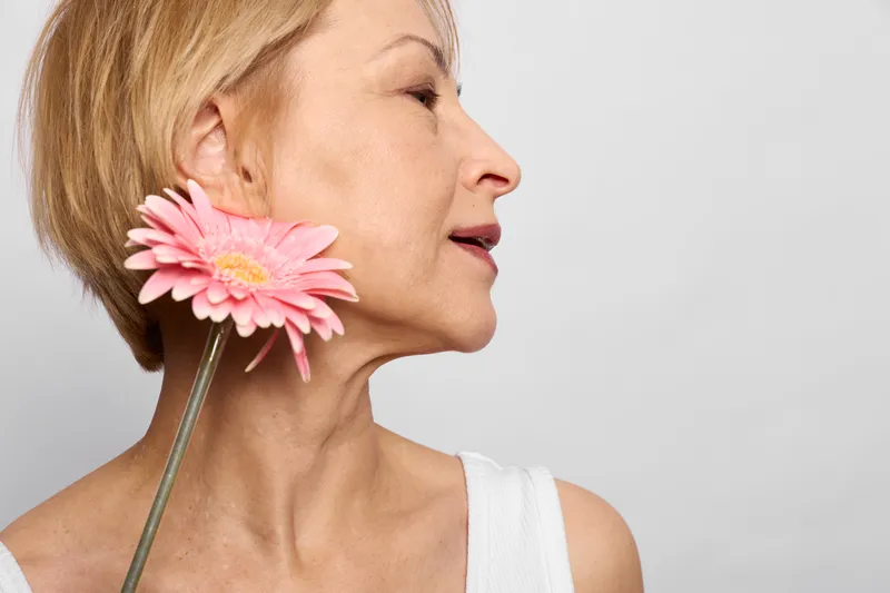 Woman with a pink flower closeup showing beauty and sensual aroma skincare, breathing beside petals on a straw, against a clean white studio background.