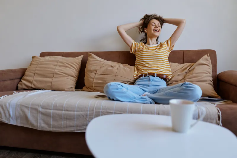 A woman sits cross legged on a cozy sofa at home, wearing a striped shirt, feeling relaxed and cheerful. Cushions surround her as natural light fills the casual living space.