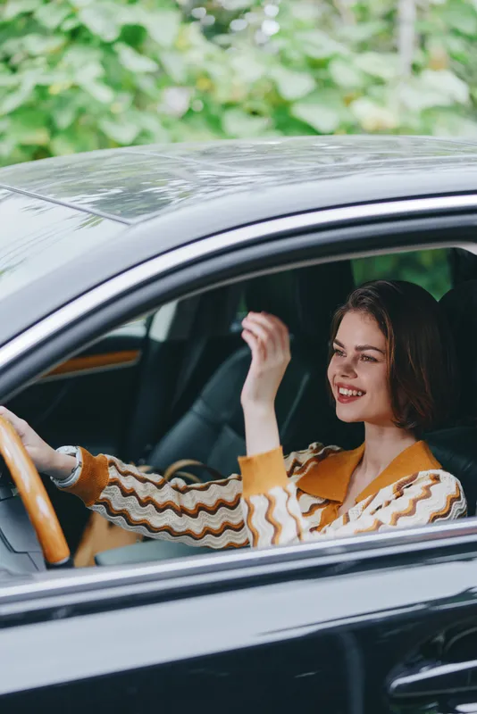 Young woman driving a car, smiling as she enjoys the ride, hands on the wheel, casual outfit, sunny day, nature softly blurred outside.