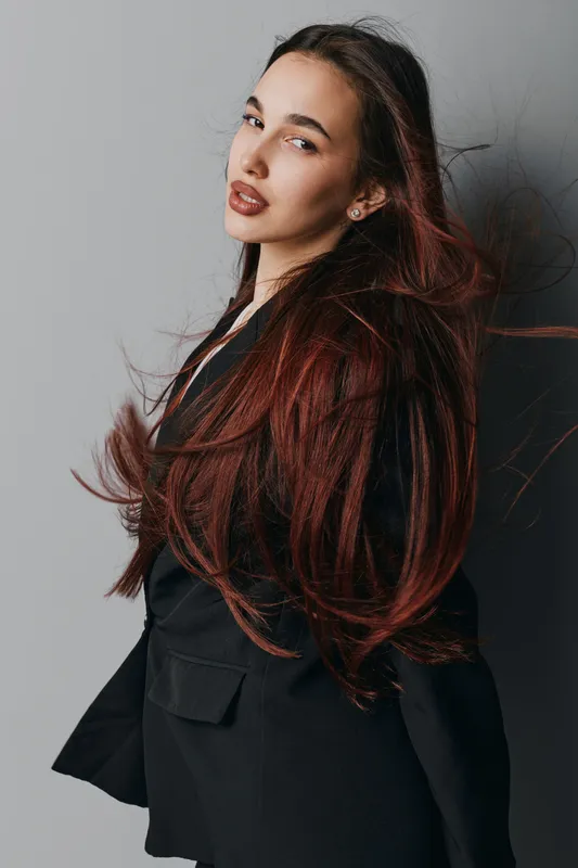 Young woman with long red hair wearing black jacket posing against gray background with hair flowing and confident expression for fashion portrait and style concept.