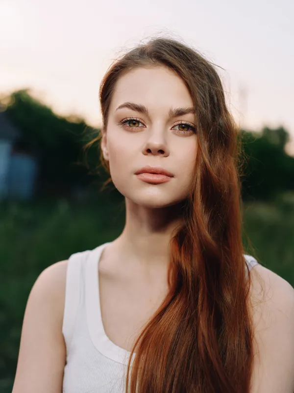 Young woman with long reddish hair in a white tank top, showcasing natural beauty and calm emotions against a blurred green background during sunset.