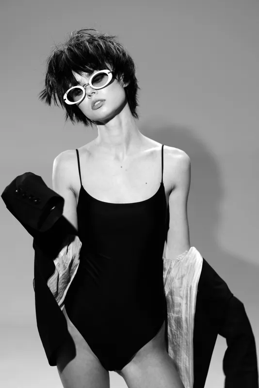 Black and white portrait of a stylish woman wearing sunglasses and a black bodysuit with minimalistic modern fashion style.
