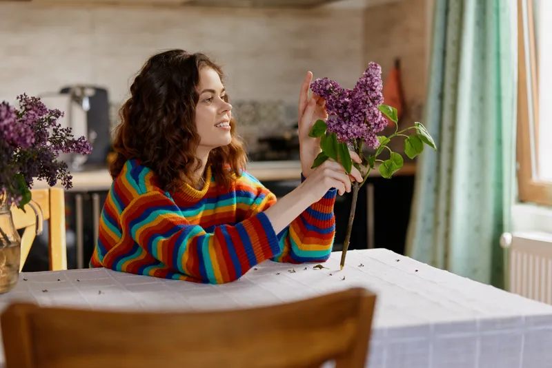 A woman in a striped colorful sweater sits at a home table, arranging lilac flowers with a gentle smile. Bright window light and a cozy kitchen in the background convey relaxed domestic moments.