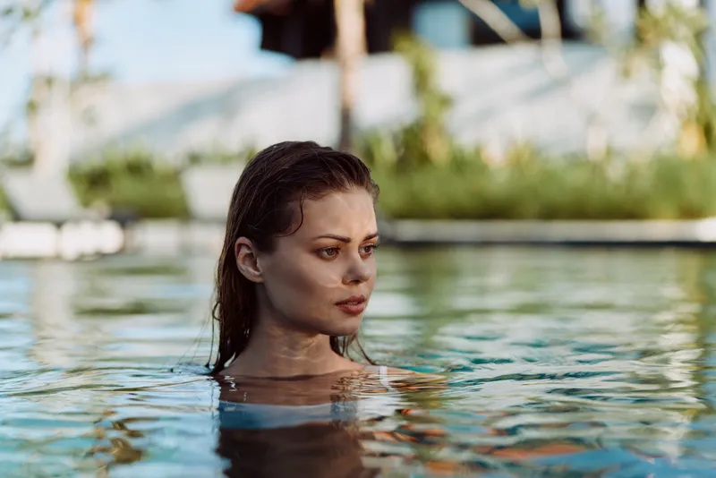 A woman calmly floats in a sunlit pool, head and shoulders above water, conveying relaxation and lifestyle leisure. Warm light, tranquil surface, and reflective water create a serene moment.