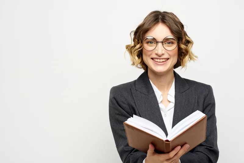 Женщина на работе с книгой в руке на светлом фоне, в классическом костюме и очках