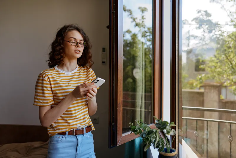 A young woman with curly hair in a striped yellow shirt stands by an open balcony window, scrolling on her smartphone in a cozy home setting.