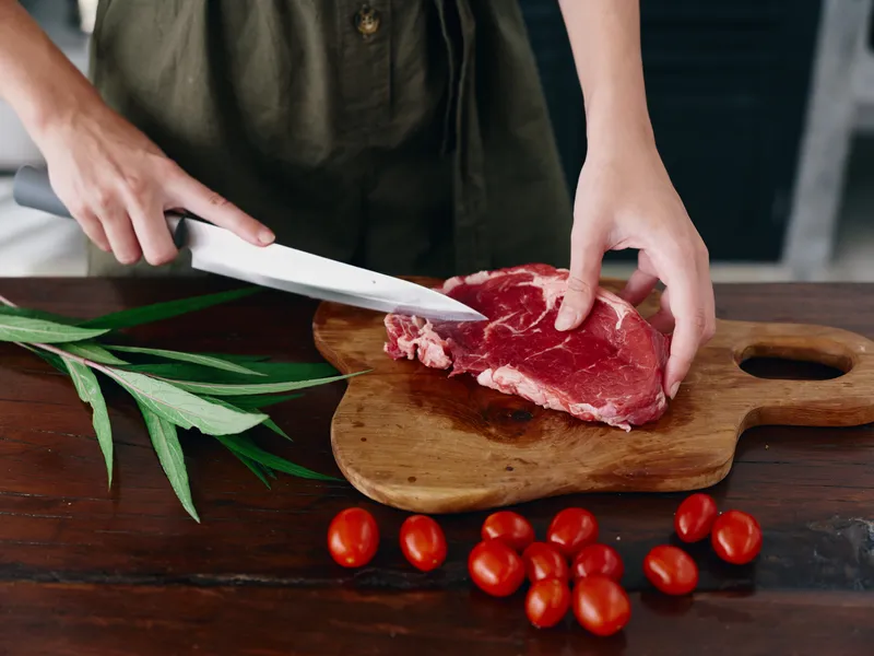 Женщина с ножом в руке нарезает свежий стейк из мяса для жарки на кухне