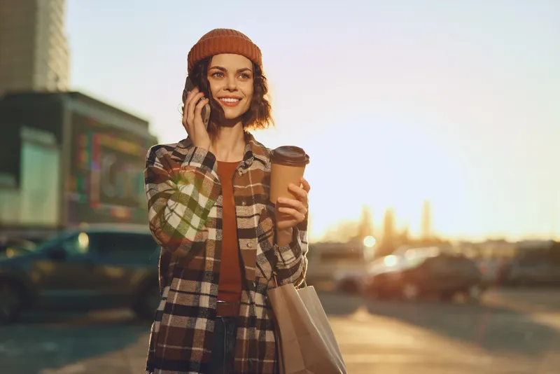 Woman coffee phone shopping urban sunlight smile portrait, holding takeaway cup and bag on city street, authenticity, candid lifestyle and golden hour glow with mindful living and emotional