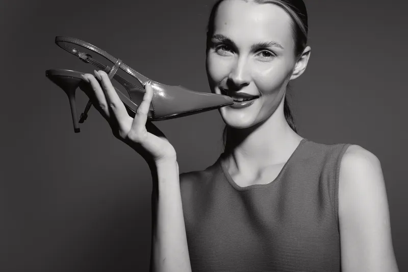 Young white woman with fair skin and blonde hair holds a high heel shoe near her lips smiling. Studio portrait in black and white with fashion and elegance concept.