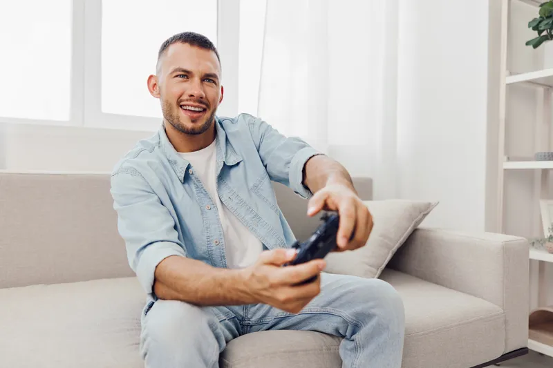 Happy man gaming at home with a modern console, showcasing enthusiasm and relaxation in a cozy living room setting filled with natural light.