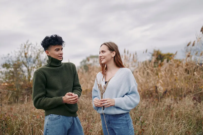 Two friends converse in a field, wearing cozy sweaters, conveying authenticity and credibility through relaxed posture and genuine smiles amid a soft autumn landscape.