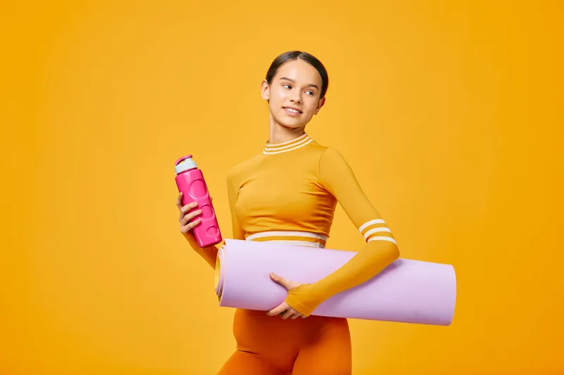 Active woman in bright orange athletic wear holds a pink water bottle and rolled yoga mat against a bold orange backdrop, showcasing fitness, sport, and healthy lifestyle.