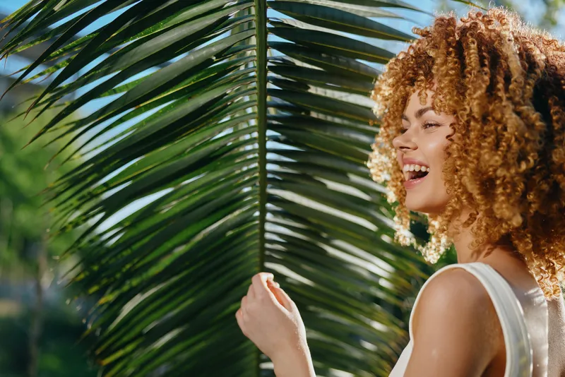 smiling woman with curly hair enjoying tropical sunlight behind large green palm leaf outdoors in summer