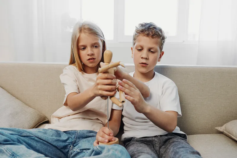 Two focused children sitting on a couch playing with a wooden mannequin toy in a bright room. Concept of childhood creativity, learning, and sibling interaction in a casual home environment.