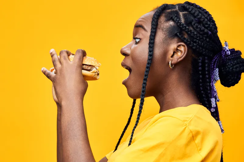 Young Black woman with braids joyfully eating a burger against a vibrant yellow background, showcasing fun, youthfulness, and delicious fast food