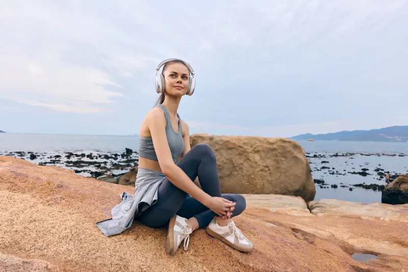 Young woman enjoying music in sportswear by the ocean, expressing relaxation, with a soft color palette of blues and earth tones, ideal for fitness and wellness concepts