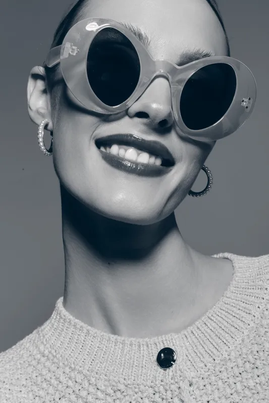Close-up of a smiling young Caucasian woman with pale skin wearing oversized sunglasses and hoop earrings. She wears a knitted sweater in a studio setting with soft lighting.