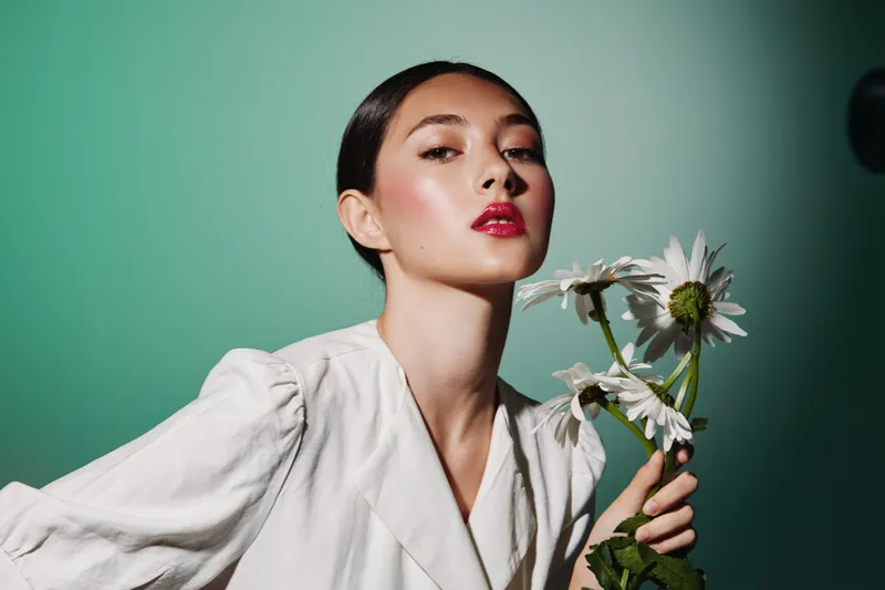 Stylish young woman with sleek hair and shiny red lips holding white daisies. Minimalist green background emphasizes natural beauty and fashion concept with creative makeup highlights.