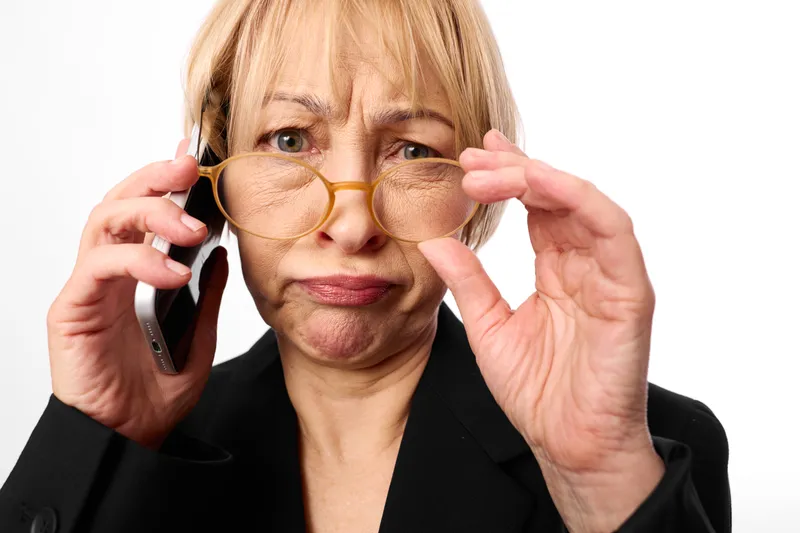 Elderly woman adjusts eyeglasses while holding a phone in a studio portrait, white background, senior vision, focused expression communication and eyewear care