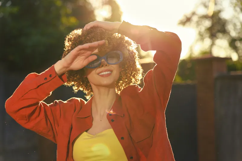 An outdoorsy woman in a red jacket and yellow top shields her eyes with sunglasses, enjoying sunlight. A candid lifestyle portrait highlighting cheerful energy, casual fashion, and bright mood