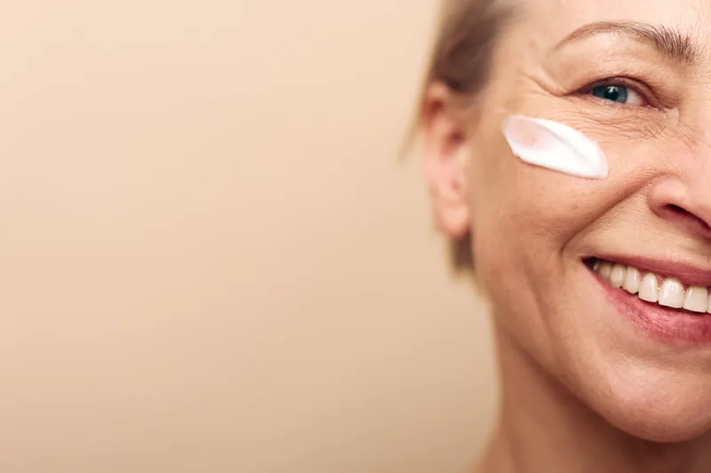 Close-up of smiling mature woman with natural skin applying white face cream on cheek on beige background. Concept for skincare, antiaging, selfcare, beauty and wellness routine.