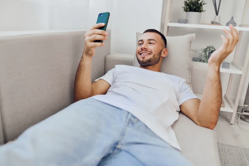 Relaxed man lounging on a couch, engaged in a fun conversation on a smartphone, creating a cozy and modern atmosphere at home, perfect for lifestyle shoots.