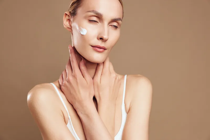 Calm portrait of a woman applying cream to her face, soft lighting, serene expression, skincare routine in progress, natural tones and relaxed posture, simple background