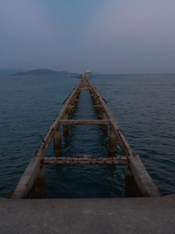 Rusty old pier extending into calm ocean waters under a cloudy sky with distant islands on the horizon.