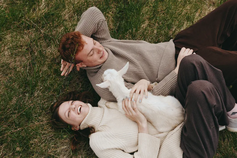Couple lying on grass outdoors with a white goat kid, sharing a tender moment and gentle touch, surrounded by natural light and calm atmosphere. The scene emphasizes lifestyle, wellbeing, and