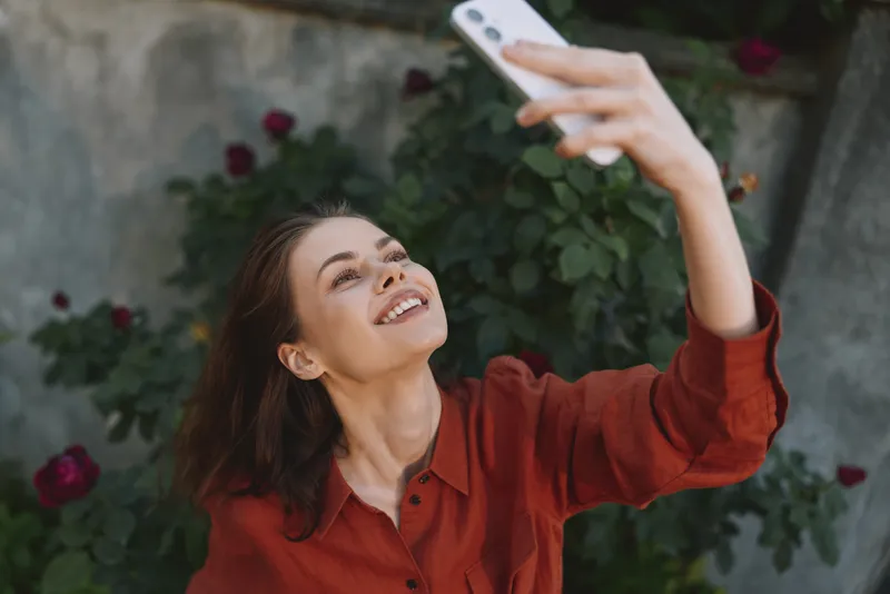 Smiling woman with shoulder length hair taking a selfie outside, wearing a red blouse, garden background. Concept of lifestyle photography, self expression, and casual moments. Soft natural light