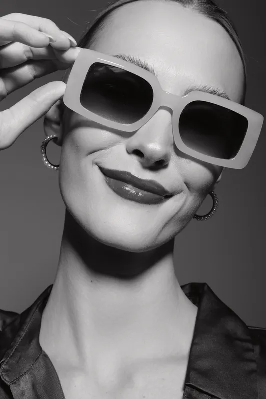 Portrait of a smiling white woman wearing large sunglasses and hoop earrings in a black shirt. Fashion and confidence concept.
