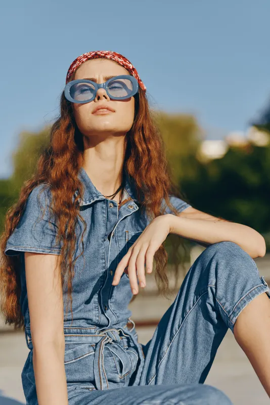 Lifestyle woman in modern boho-western denim street style wearing blue sunglasses and a red headband with wavy hair under warm natural sunlight film color tone.