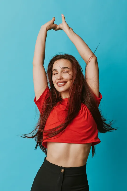 Young woman with long hair smiling and dancing with raised arms wearing red crop top and black pants against blue background. Happy girl enjoying movement and freedom indoors.
