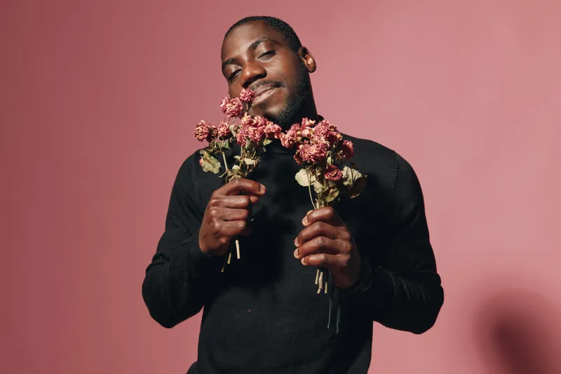 Creative conceptual portrait of African man holding dried flowers with closed eyes and smile in visual style. Black male model expressing emotion with pink background in studio.
