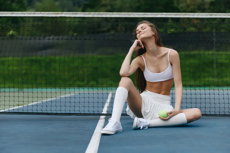 Stylish athletic woman sitting on tennis court, wearing white sportswear, showing joy and relaxation, representing active lifestyle and seasonal sports concept