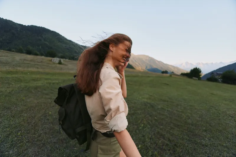 Young woman with a backpack walking through a meadow beneath mountains, showcasing outdoor lifestyle and travel with vibrant nature all around.