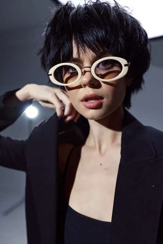 Stylish woman wearing oversized round sunglasses and a black blazer, posing with confident expression and short dark hair.