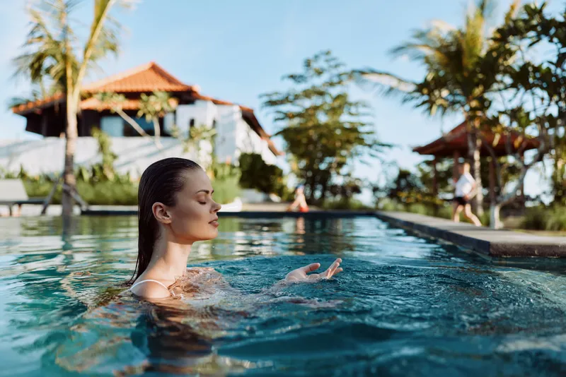 Woman relaxing in a sunny pool scene, calm water shimmering under a tropical resort backdrop, leisure lifestyle by the pool, outdoor vacation vibe, serene mood, casual swim, peaceful moment by the