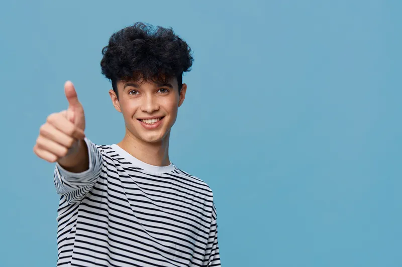 Smiling young man in black and white striped shirt shows thumbs up gesture with friendly expression on blue background, symbolizing positivity and approval.