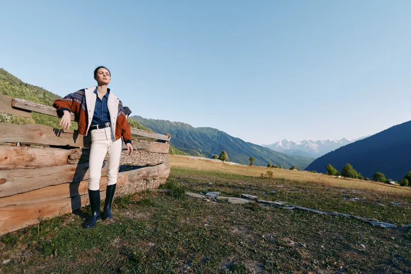 Man leaning on wooden fence in mountains meadow wearing stylish coat and boots, countryside portrait with wide sky and scenic landscape, fashion pose and relaxed outdoors vibe.