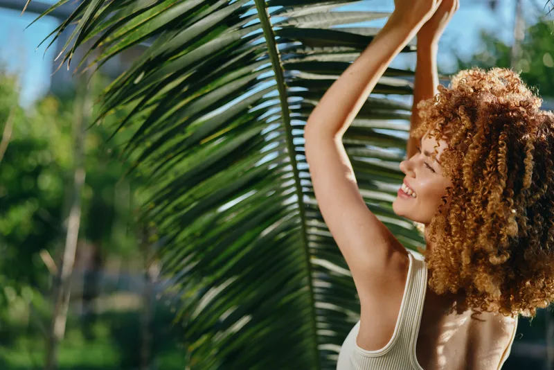smiling woman with curly hair enjoying nature, tropical leaf background, bright daylight, joyful mood, summer outdoors, natural beauty portrait